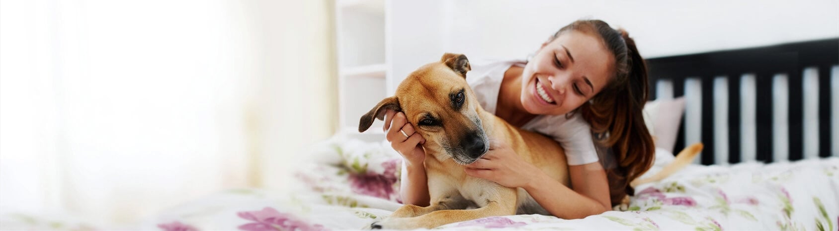 A smiling woman embracing a light brown dog on a bed with floral sheets.
