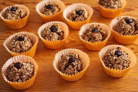 A dozen oatmeal muffins with blueberries on top sit in yellow cupcake liners on a wooden surface.