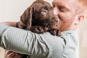 A man with red hair holds a brown puppy, looking at it with a smile.