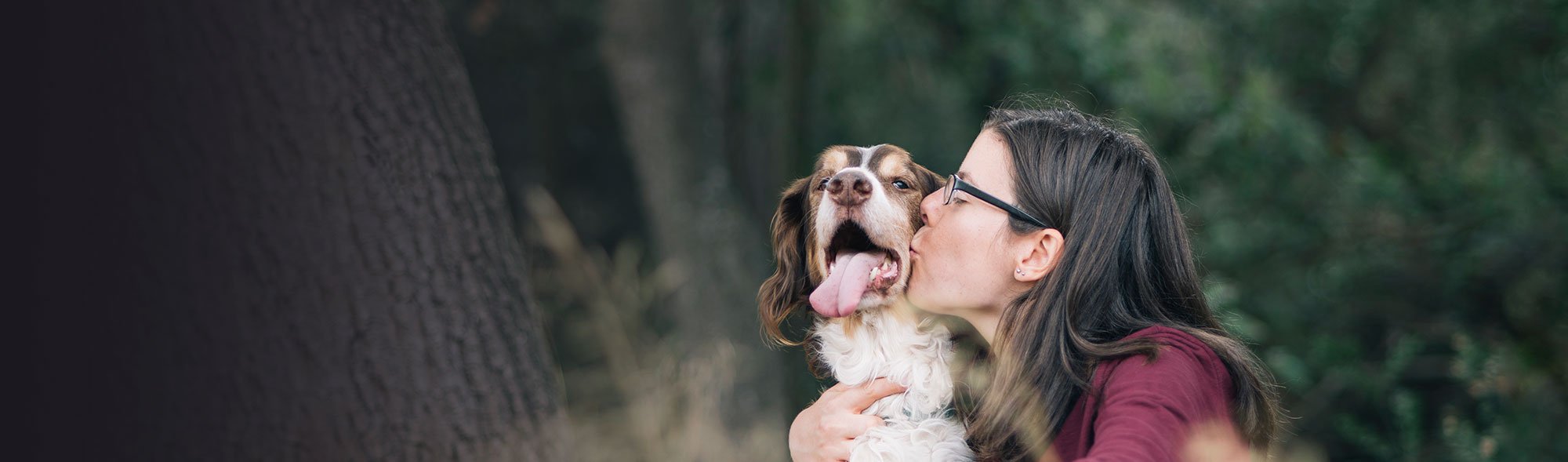 A woman with glasses kisses a happy brown and white dog with its tongue out.