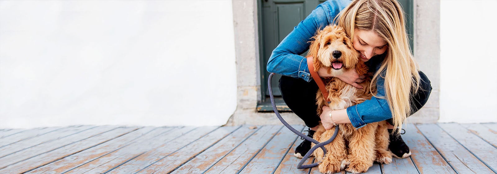 A blonde woman in a denim jacket crouches to hug a fluffy golden dog with a leash.