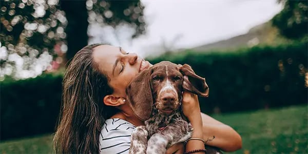 A woman with long hair and closed eyes hugs a brown and white spotted dog.