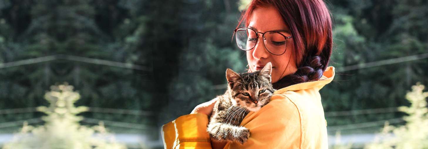 A woman with glasses and a braid holds a tabby cat on her shoulder, looking at it lovingly.