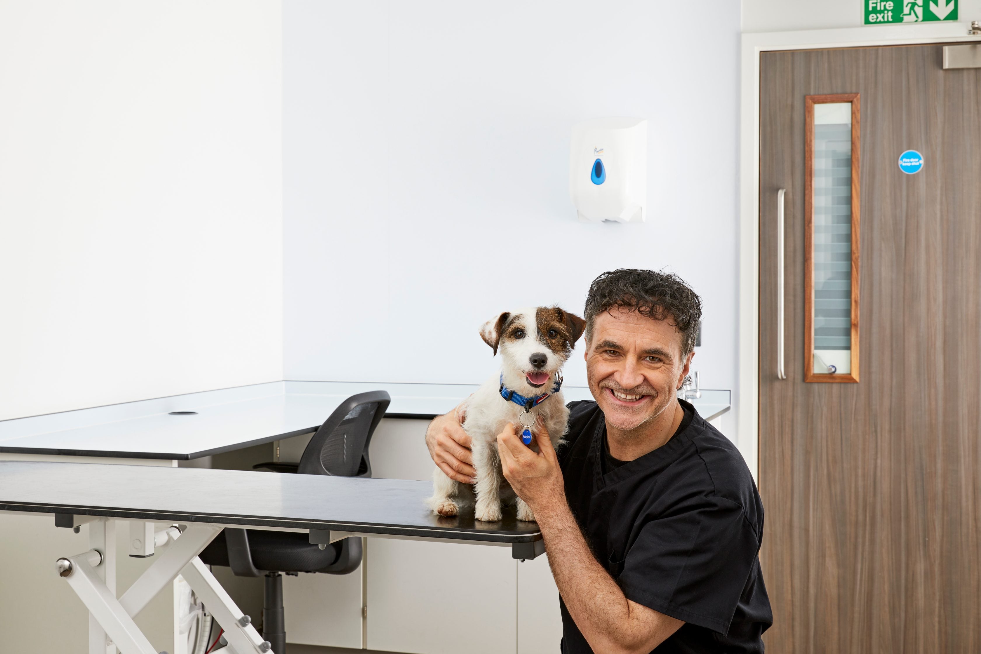 A smiling veterinarian in black scrubs holds a brown and white terrier on an examination table.